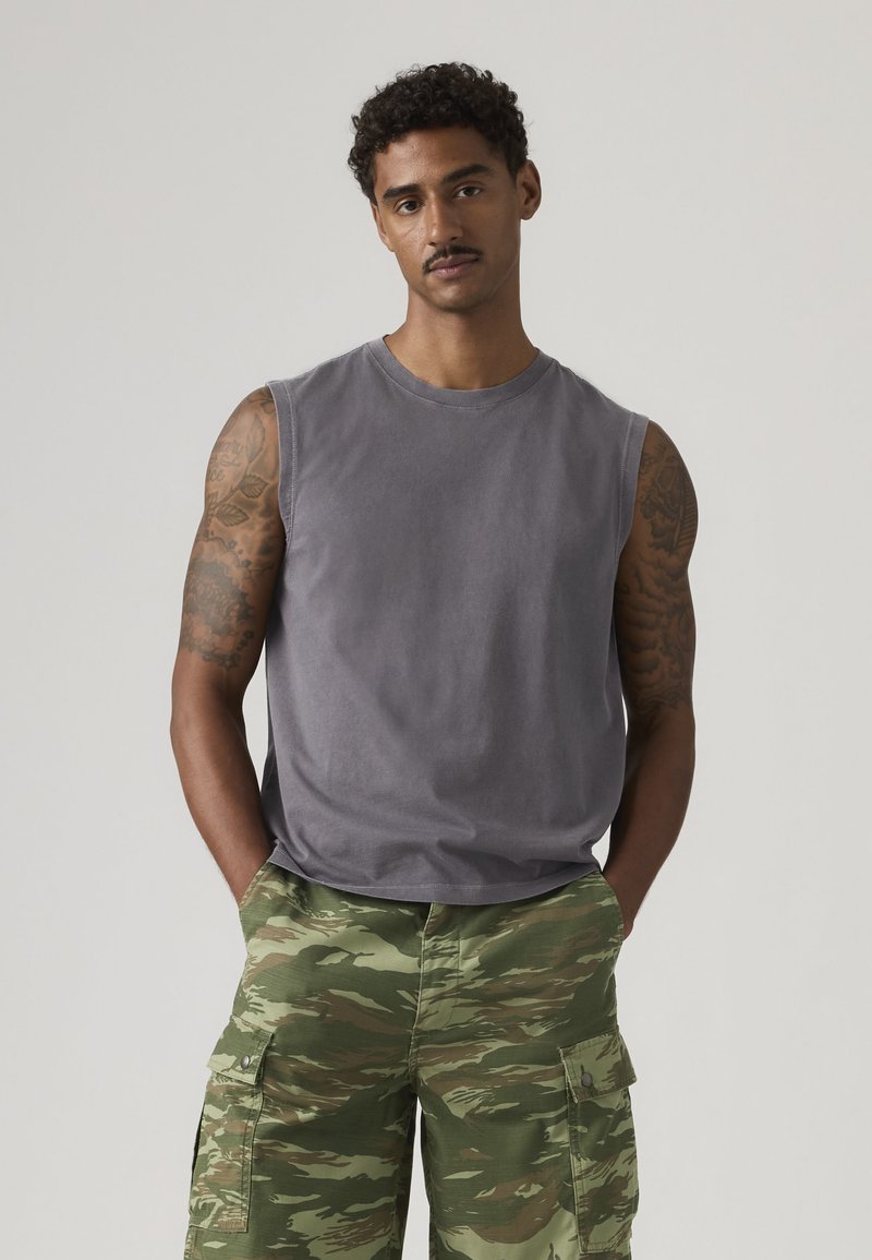Homme tatoué portant un débardeur gris et un short cargo camouflage vert, debout les mains dans les poches sur un fond uni.