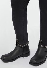Botas negras hasta el tobillo hechas de cuero suave, con tachuelas plateadas y acentos de estrellas a lo largo del puño, combinadas con jeans negros ajustados.