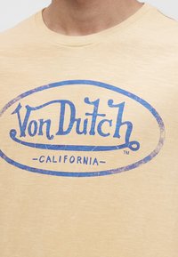 Personne portant une chemise beige avec un logo bleu "Von Dutch California" usé imprimé sur la poitrine.