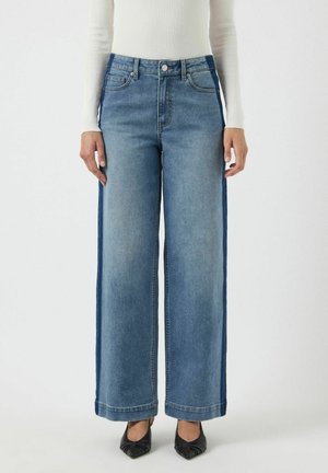 Pantaloni in denim a gamba larga con lavaggio medio, pannelli laterali più scuri a contrasto e orli rifiniti. Abbinati a scarpe nere con punta affusolata.