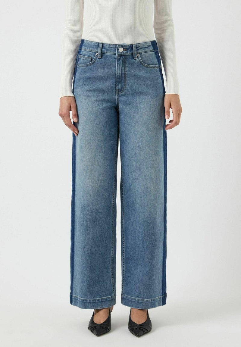 Pantalones de denim de pierna ancha con un lavado medio, paneles laterales oscuros contrastantes y dobladillos en los tobillos. Combinados con zapatos negros de punta afilada.