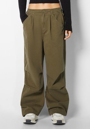 Personne portant un pantalon ample vert olive avec une taille élastique, un crop top noir à manches longues et des baskets blanches, debout devant un fond uni.