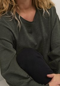 Felpa verde scuro con una texture morbida, maniche larghe e scollo arrotondato. Una persona è accovacciata indossando pantaloni neri.