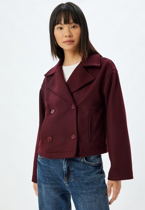 Bordeaux dubbelzijdig jasje van zachte stof met gesneden kraag, voorzakken en rode knopen, gecombineerd met blauwe jeans.