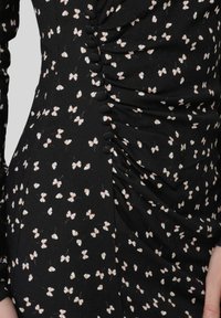 Zwarte stoffen jurk met kleine, roze vlinderprints. Heeft geplooide details aan de zijkant en decoratieve knopen langs de naad.