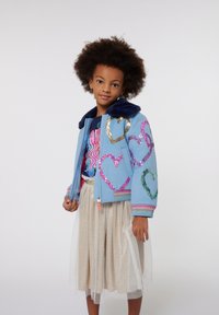 Giacca bomber azzurra chiara con colletto navy, caratterizzata da design a cuore in paillettes multicolori. Abbinata a una gonnellina in tulle beige scintillante.