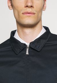 Črna zip-jakna z obrnjenim ovratnikom. Ima kovinski zadrgo in gladko, teksturirano tkanino. Preprost, minimalističen dizajn.