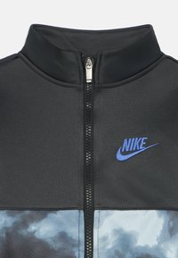 Casaco preto com fecho zip e gola alta. Apresenta um logotipo da Nike em azul e um desenho inferior com um padrão nublado em azul claro e cinza. Textura suave.