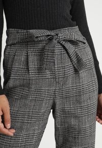 Šedé kostkované kalhoty s vysokým pasem a mašlí v pase, vyrobené z texturované tkaniny a s faldy pro zvýšení detailu a tvaru.