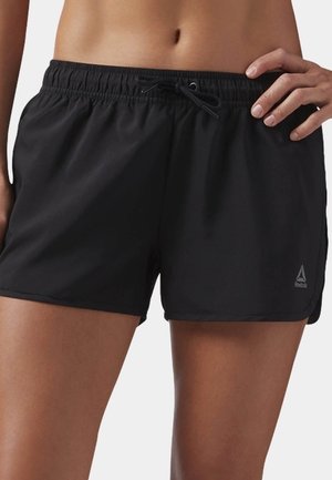 Nærbillede af en person iført sorte Reebok sportsshorts med elastisk taljebånd og snøre, hånden hvilende på hoften.