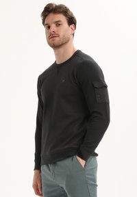 Zwarte sweatshirt met lange mouwen en geribbelde manchetten, voorzien van een zak op de linker mouw. Glad textuur en minimalistisch ontwerp.
