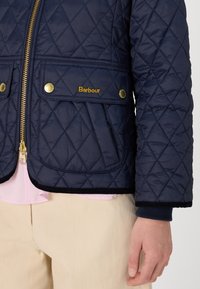 Navy quilted jacket with gold zipper and accents, two front pockets, and embroidered logo. Features ribbed cuffs and hem detail.