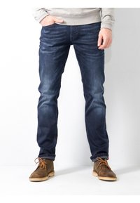 Vaqueros de mezclilla azul oscuro con un diseño ajustado, que cuentan con un ligero desgaste, estilo de cinco bolsillos y botas de gamuza marrón desgastadas con cordones.