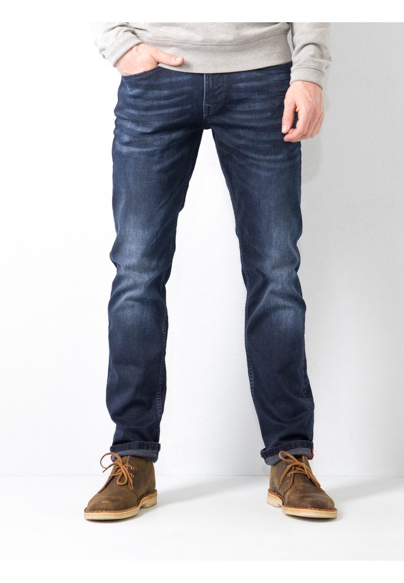 Vaqueros de mezclilla azul oscuro con un diseño ajustado, que cuentan con un ligero desgaste, estilo de cinco bolsillos y botas de gamuza marrón desgastadas con cordones.