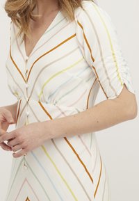 Randig klänning med korta, rynkade ärmar. Färgerna inkluderar vit, orange, blå, rosa och grön. Har en v-ringning och knäppning.