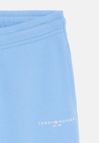 Gaiši zilie sporta bikses, kas izgatavotas no mīksta auduma, ar baltu logotipu un tekstu "TOMMY HILFIGER EST. 1985" priekšpusē.