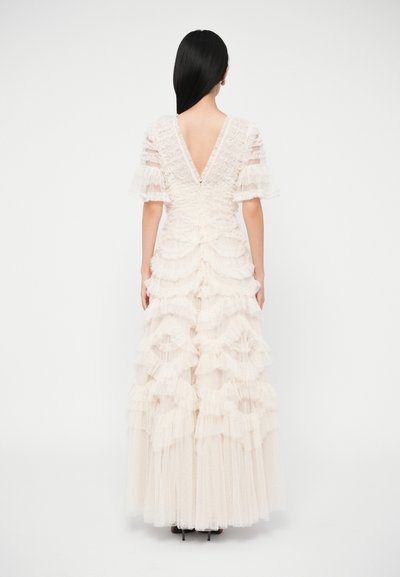 Kobieta o długich czarnych włosach, ubrana w kremową suknię sięgającą podłogi z warstwowymi falbanami i głębokim dekoltem na plecach, stojąca na białym tle.