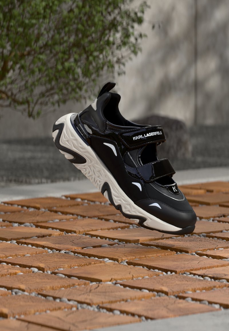Μαύρο και λευκό αθλητικό παπούτσι Karl Lagerfeld με λουριά βέλκρο να αιωρείται πάνω από ξύλινο και χαλίκι έδαφος σε εξωτερικό χώρο κοντά σε πράσινη φυτοκομία.