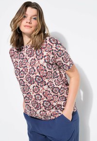 Blümchenmuster-T-Shirt in Creme, mit roten und blauen Blumen, kurzen Ärmeln, Rundhalsausschnitt und lässiger Passform. Kombiniert mit marineblauen Hosen.