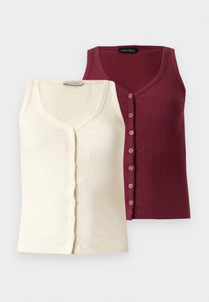 Femme debout, main dans la poche, portant un haut bordeaux sans manches à boutons, un jean bleu à jambes larges et des chaussures noires à talons.