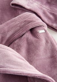 Weicher, gerippter Stoff in zartem Rosa. Enthält ein Metalletikett mit dem Wort "DOWNTIME." Verfügt über eine glatte Textur und ein mehrschichtiges Design.