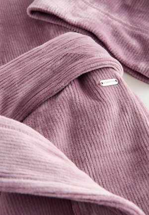 Blødt, ribbet stof i lyserød. Inkluderer et metalmærke med ordet "DOWNTIME." Har en glat tekstur og lagdelt design.