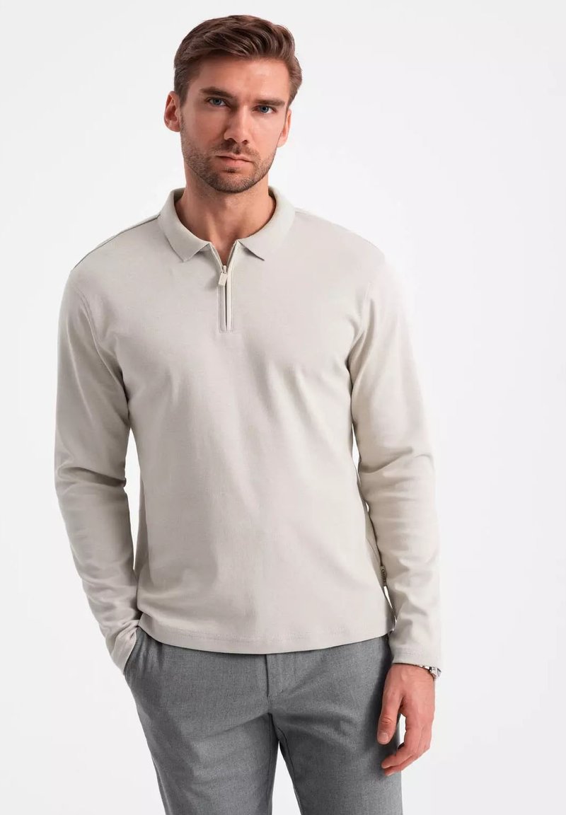 Hellbeige Langarm-Polohemd mit Reißverschlusskragen, aus weichem Stoff, getragen mit hellgrauen Hosen, die einen lässigen Schnitt haben.