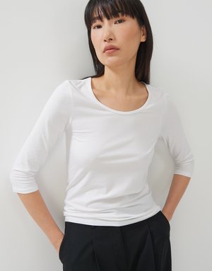 Top bianco a maniche lunghe, aderente, con scollatura rotonda, realizzato in tessuto morbido, abbinato a pantaloni neri, mani nelle tasche.