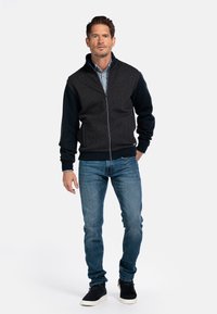 Heren zip-up jack met een grijze textuur op het lichaam en zwarte mouwen, gecombineerd met blauwe jeans en donkere sneakers. Gladde, casual uitstraling.