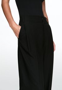 Pantalon noir à texture lisse, doté d'une fermeture éclair et de poches latérales, conçu avec une coupe décontractée et un devant plissé.