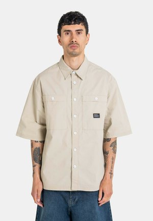 Mann mit kurzen dunklen Haaren und Tätowierungen trägt ein beiges kurzärmeliges Hemd mit zwei Brusttaschen und blaue Jeans-Shorts.
