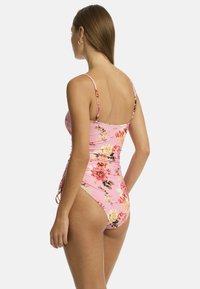 Une jeune femme se tient de dos face au spectateur, portant un maillot de bain floral rose à fines bretelles, mettant en valeur le motif floral coloré.
