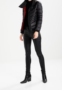 Mustat puffatakki punaisella vuorilla; muotoiltu malli. Mustat mokkatakki housut kiiltävällä pinnalla. Mustat korolliset nilkkasaappaat.