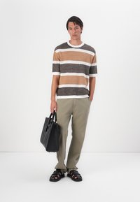 Maglione a righe in beige, marrone e bianco, abbinato a pantaloni verde oliva. L'uomo tiene una borsa a tracolla nera e indossa sandali neri.