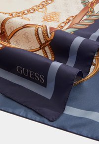 Foulard en soie avec un motif chaîne en tons dorés et bruns, bordé de bleu marine, affichant le texte "GUESS" en blanc sur un fond sombre.