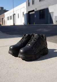 Paire de baskets noires épaisses avec des semelles épaisses posées sur un trottoir en béton dans un cadre urbain.
