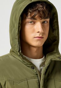 Chaqueta con capucha de color verde oliva, con cierre de cremallera y broche a presión, que presenta una textura suave y un discreto detalle de costuras. Camisa blanca debajo.
