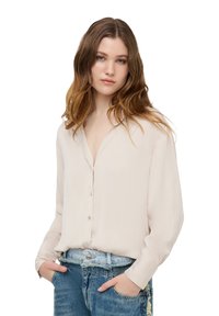 Camicia button-up beige chiaro con colletto, maniche lunghe e una texture liscia, abbinata a jeans blu strappati.