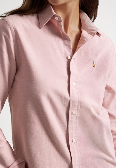 Svetloružová košeľa na gombíky z bavlny. Má špicatý golier, biele gombíky a malú vyšívanú značku na ľavej strane hrudníka.