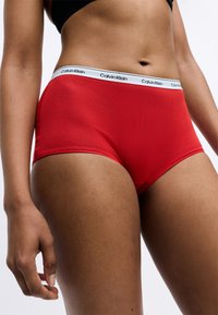 Piros pamut bugyi, fehér derékrésszel, amelyen a "Calvin Klein" logó található. Az anyag sima tapintású és testhez álló dizájnnal rendelkezik.