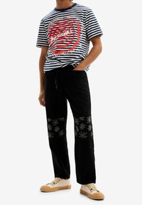 Pasiasty niebiesko-biały t-shirt z czerwonym grafiką, czarne spodnie z haftowanymi kwiatowymi wzorami oraz beżowe buty z fakturowanymi podeszwami.