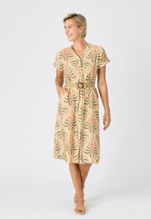 Femme souriante, portant une robe midi beige à motifs floraux, manches courtes, boutonnée devant, taille cintrée avec une ceinture, et des talons beige à bride à la cheville, debout sur fond blanc.