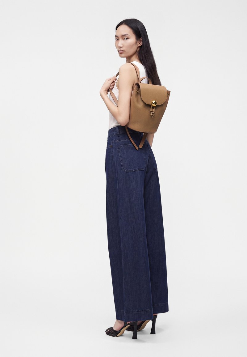 Femme aux longs cheveux noirs portant un haut blanc sans manches, un jean bleu à jambes larges, des talons noirs et un sac à dos en cuir marron sur une épaule.