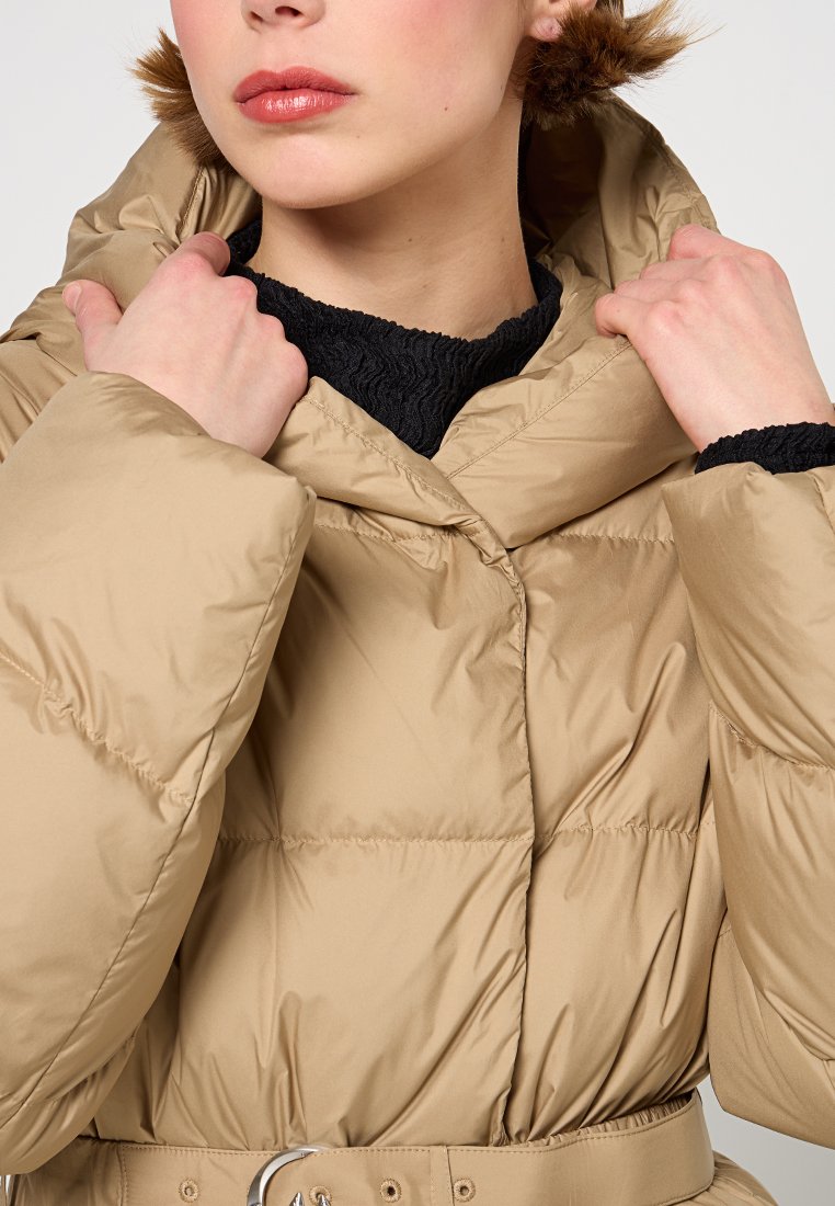 Giacca puffer beige con colletto alto e cintura. Presenta una texture liscia e lucida e un design trapuntato, indossata sopra un top nero con texture.