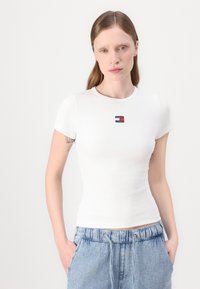 Hvit ribbet kortermet topp med en liten rektangulær Tommy Jeans-logo i svart, rødt og blått på brystet. Glatt tekstur, tettsittende design.