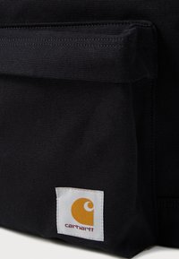 Acercamiento de una bolsa de tela negra con solapa cosida y parche del logo de Carhartt en amarillo y blanco en la esquina inferior.