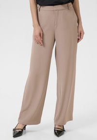 Beige wijde broek gemaakt van zachte stof, met een medium taille en schone plooien aan de voorkant, gecombineerd met zwarte puntige hakken.