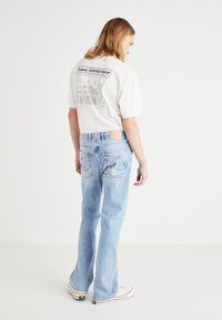 Svetlo modri denim hlače s sproščenim krojem, z označeno zadnjo nalepko in značilnimi ukrivljenimi zadnjimi žepi. V kombinaciji z belo majico s potiskom.