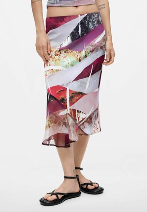 Donna con gonna midi colorata a motivo astratto con orlo arricciato e sandali neri con cinturino alla caviglia, in piedi su sfondo bianco semplice.