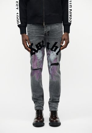 Homme portant un jean gris usé avec des textes gothiques noirs et des motifs d'ange violets, un sweat à capuche noir et des bottes à lacets marron foncé.